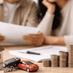 Couple reviewing financial documents at a table with house model, car keys, and stacked coins, planning a loan for major purchases